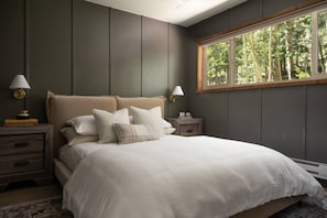 Un dormitorio con una cama grande, dos mesitas de noche, una lámpara sobre cada mesita y una ventana con vistas a los árboles.