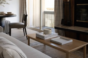 Un salon moderne avec un canapé, une table basse en bois sur laquelle sont posés des livres et une bougie, un fauteuil et une vue sur l’extérieur grâce à une grande fenêtre.