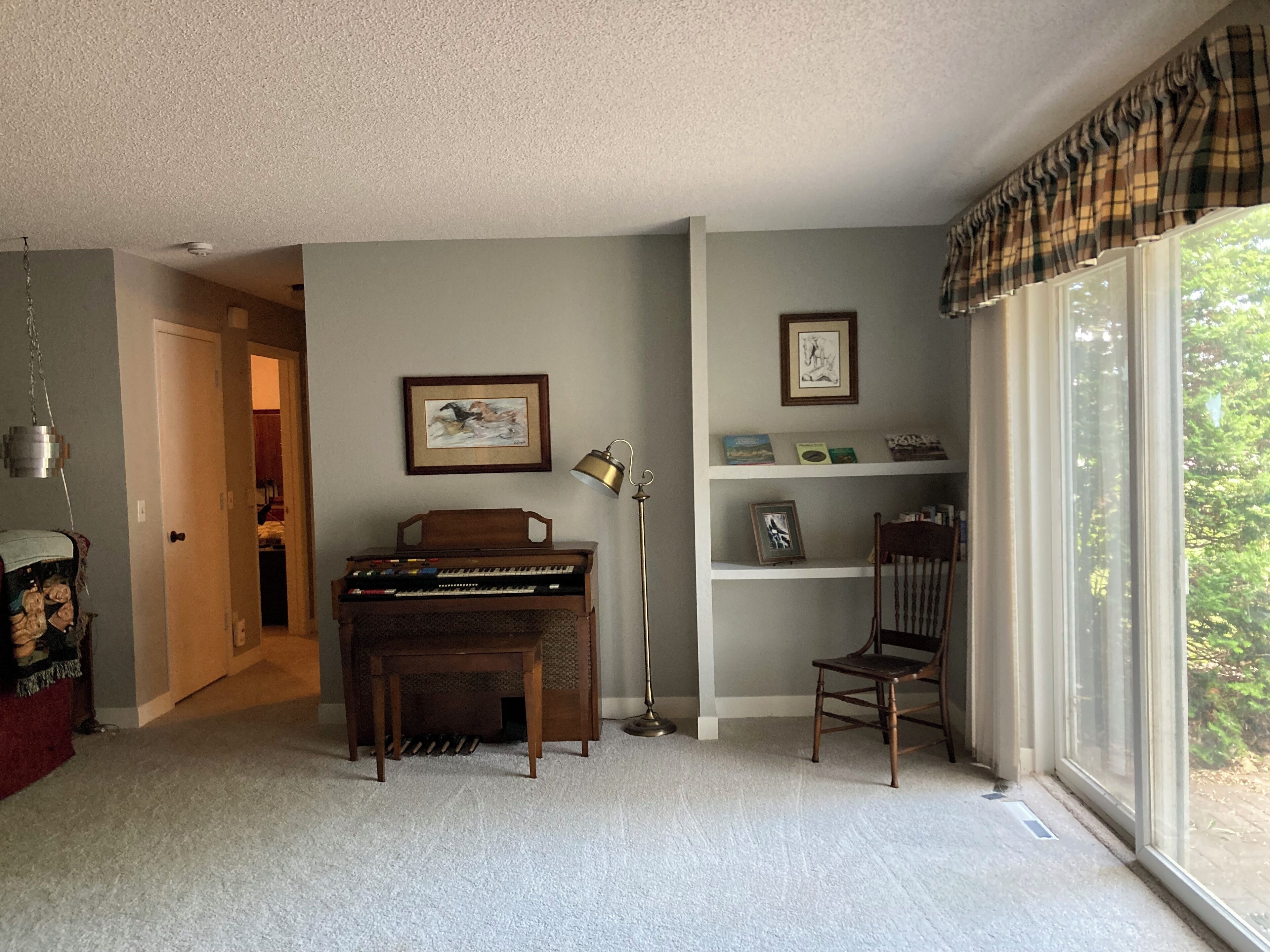 Ein Wohnzimmer mit Klavier, einem Sessel, einem Bücherregal und einem Fenster mit Vorhängen.