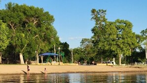 A calm river with people on the shore, a boat, and a park with trees and a blue-roofed structure. - John & Anne`s Place LLC (Litchfield)