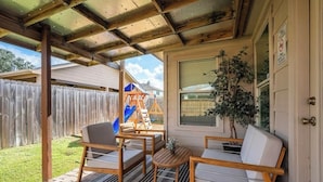 Une terrasse couverte, aménagée avec des meubles en bois, une petite table et une fenêtre donnant sur une cour arrière où se trouve une aire de jeux.