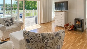 A spacious living room with a wooden floor, a white sofa, a patterned armchair, a TV, a fireplace, and a view of a deck and greenery outside.