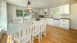 A modern kitchen with white cabinetry, a wooden dining table with chairs, a chandelier, and a view of the outdoors through large windows.