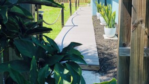 Una terraza de madera que lleva a una casa con fachada azul claro. Hay plantas en macetas a lo largo de la terraza y una valla de madera al fondo.