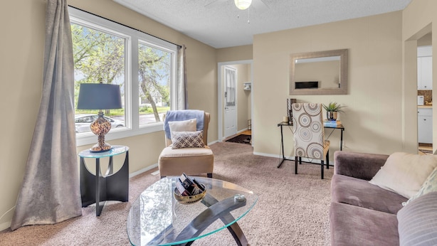A living room with a glass coffee table, a sofa, a chair, a TV, and a ceiling fan. - Mighty Manor (North Battleford)