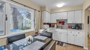 A modern kitchen with white cabinets, a dining table with chairs, and a view of the outside through a large window. - Mighty Manor (North Battleford)