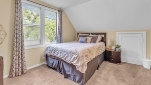 A bedroom with a bed, a nightstand, a dresser, a window with curtains, and a door. - Mighty Manor (North Battleford)