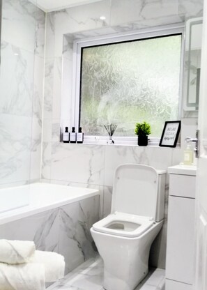 Un baño moderno con piso de mármol, una bañera blanca, un inodoro y un ventanal con diseño de vidrio esmerilado.