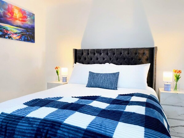 A hotel room with a large bed, a tufted headboard, white pillows, a blue and white checkered blanket, and a painting on the wall. - Newly Renovated, Clean and Spacious home close to Anfield and Everton Stadium (Liverpool)