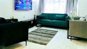 A living room with a large flat-screen TV mounted on the wall, a green sofa, a black armchair, a patterned rug, and a small table with a plant.