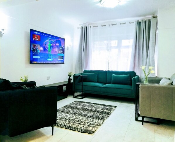 Un salón con un televisor de pantalla plana grande montado en la pared, un sofá verde, un sillón negro, una alfombra estampada y una pequeña mesa con una planta.