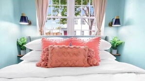 Een net opgemaakt bed met roze en witte kussens, twee wandlampen en een raam met gordijnen.