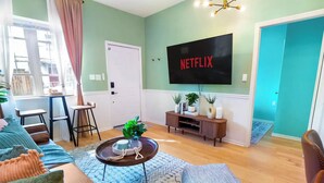 Een moderne woonkamer met een flatscreen televisie aan de muur, een bijzettafeltje met decoratieve accessoires, een bank en een kamerkleed met dessin.