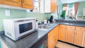 Een keuken met een magnetron, een dubbele spoelbak, houten kastjes en een raam met gordijnen.