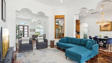 A spacious living room with a large teal sectional sofa, a dark armchair, and a TV on a stand. There is a dining area with a table and chairs, and a chandelier above.