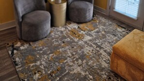 Una habitación con dos sillones grises, una mesita redonda con lámpara, una alfombra estampada y un ventanal con persianas.