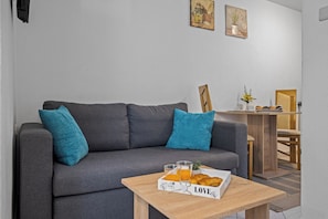 Un salon moderne avec un canapé gris, des coussins turquoise, une table basse en bois sur laquelle sont disposés des éléments pour le petit-déjeuner, et un coin repas avec une chaise et une table en bois.