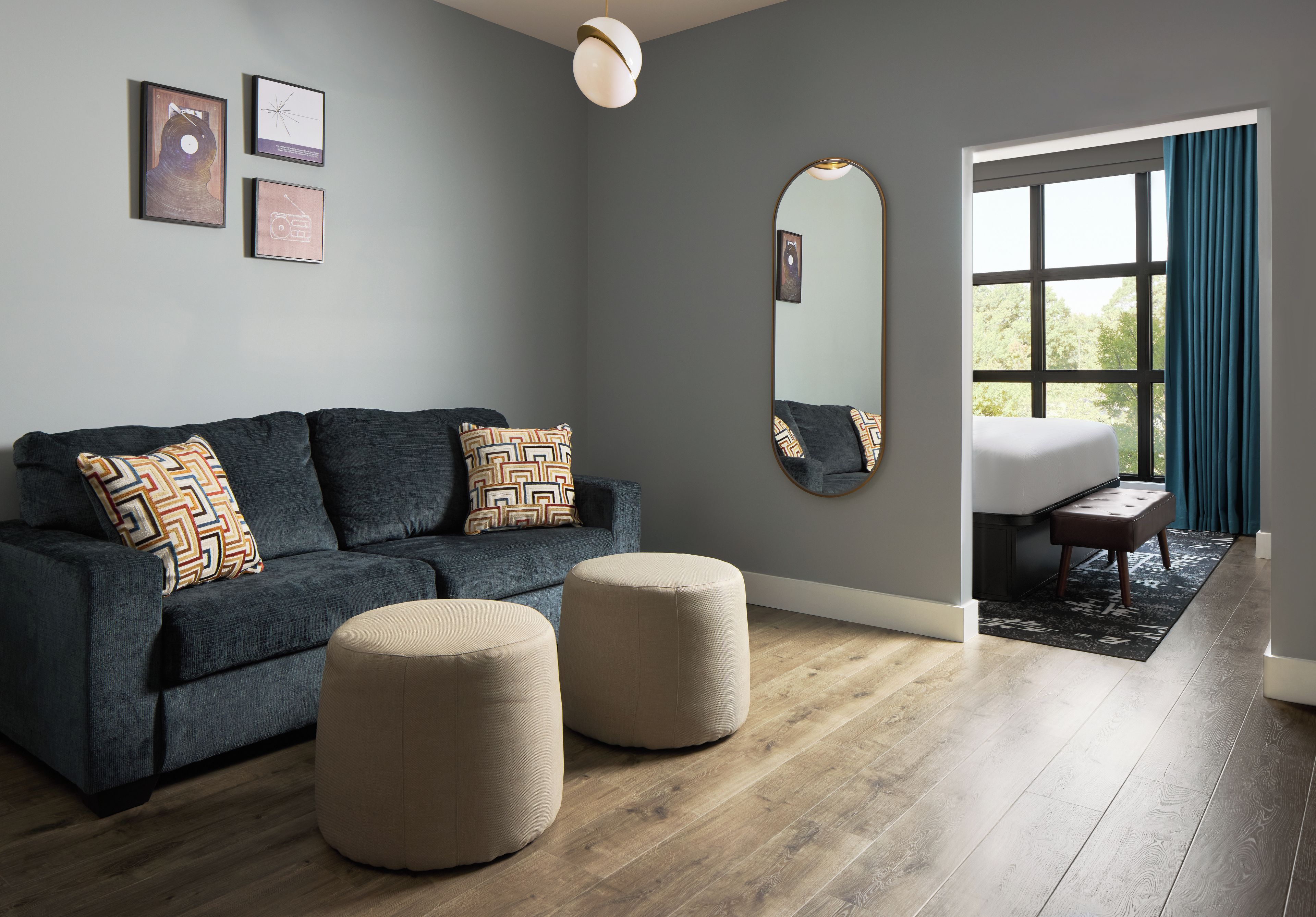 A modern living room with a grey sofa, two ottomans, a large mirror, and a view through a window with curtains.
