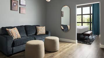 A modern living room with a grey sofa, two ottomans, a large mirror, and a view through a window with curtains.