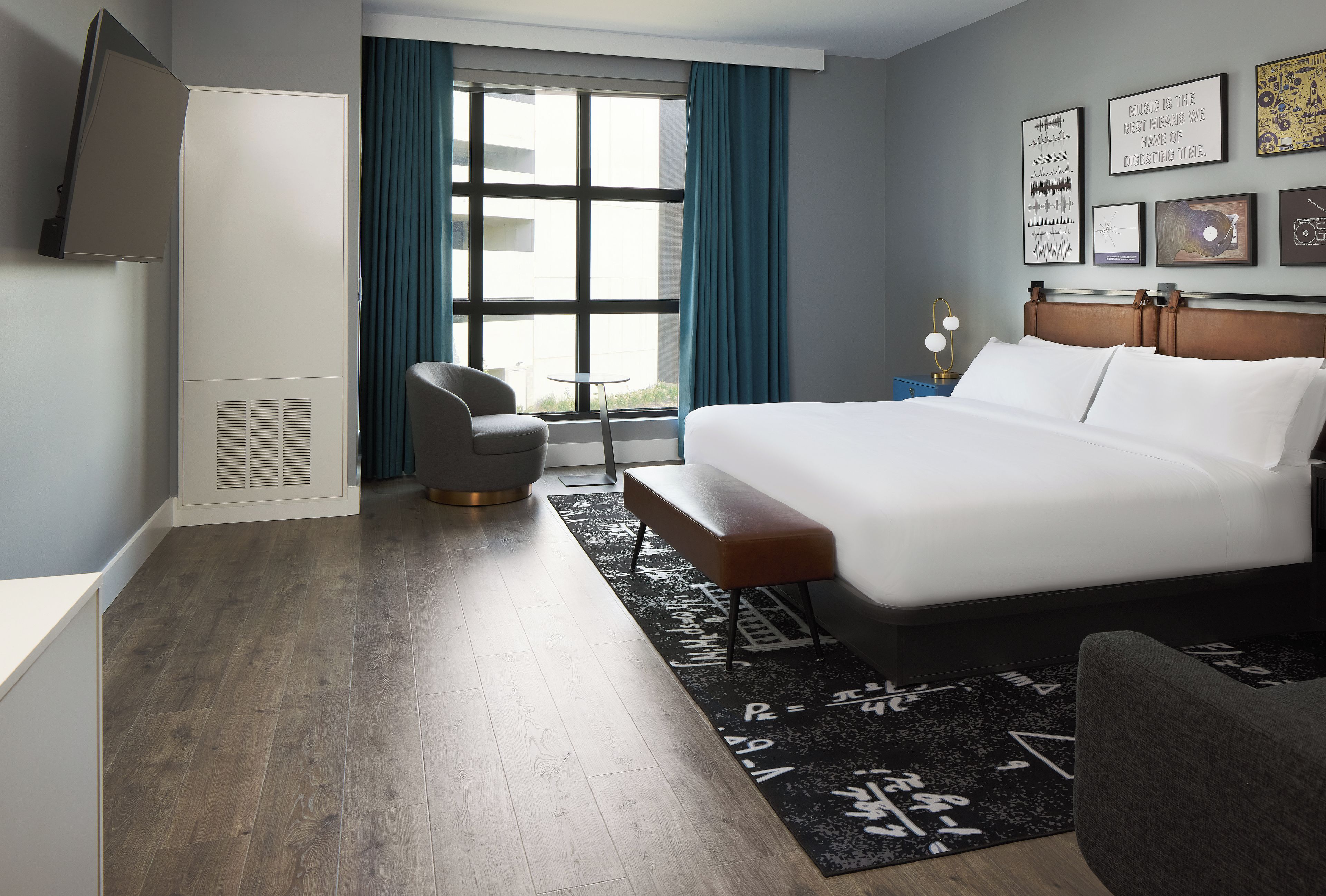 Une chambre d’hôtel avec un lit, une chaise, un bureau, une fenêtre avec des rideaux et des œuvres d’art murales.