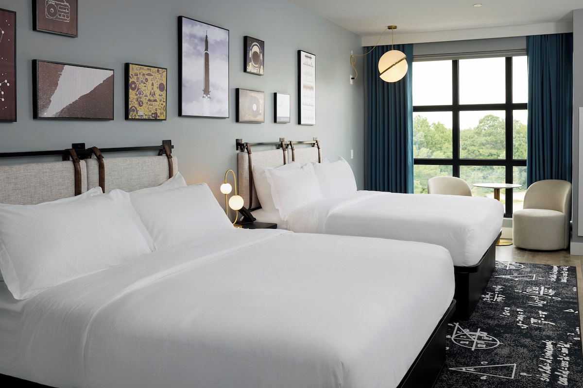 A hotel room with two beds, a nightstand, a lamp, and a view of the outdoors.