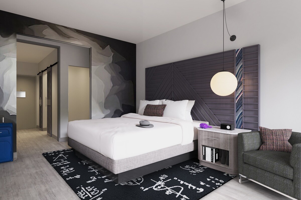 A modern bedroom with a large bed, a bedside table with a lamp, a chair, and a bookshelf.