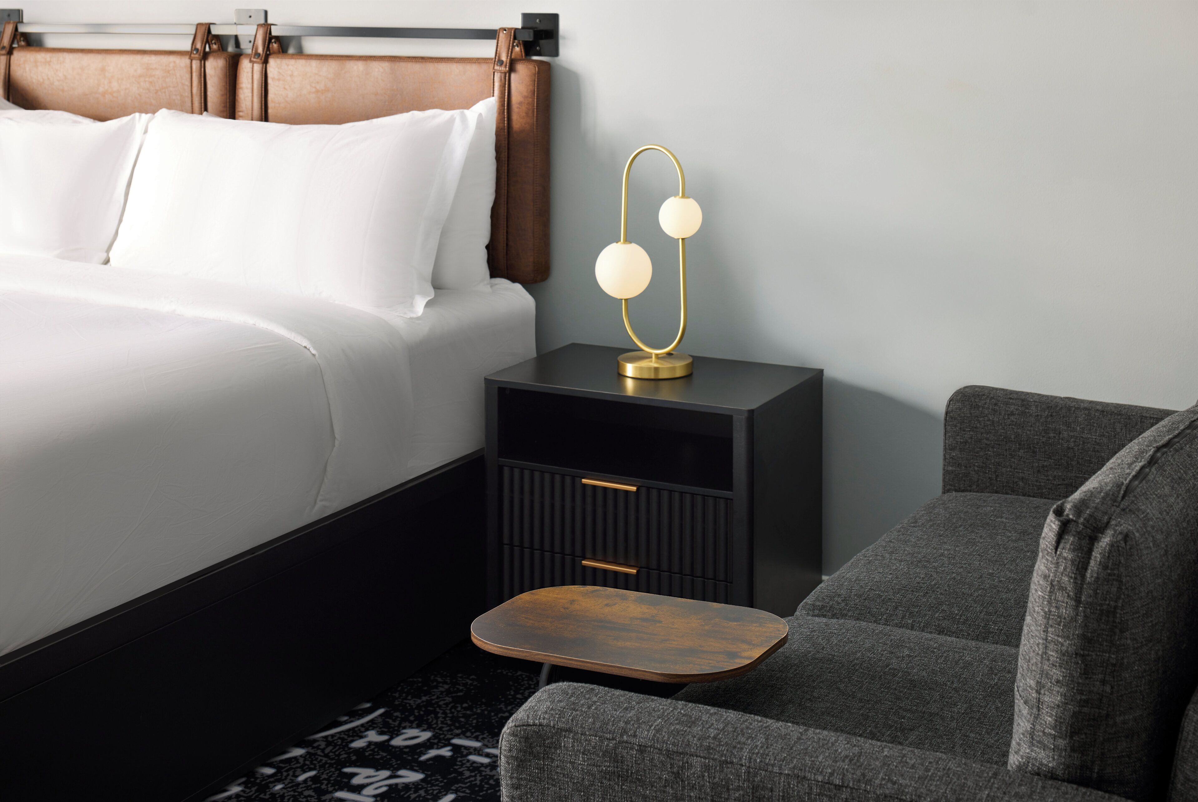 A neatly arranged hotel room with a bed, bedside table, lamp, and a small wooden stool.