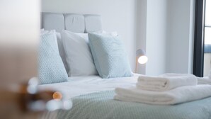 A neatly made bed with a light green bedspread and white pillows, a bedside lamp, and a window with daylight.