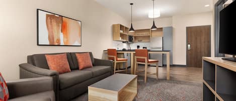 A modern living room with a sofa, coffee table, and TV stand. There is a kitchen area with cabinets and a microwave.