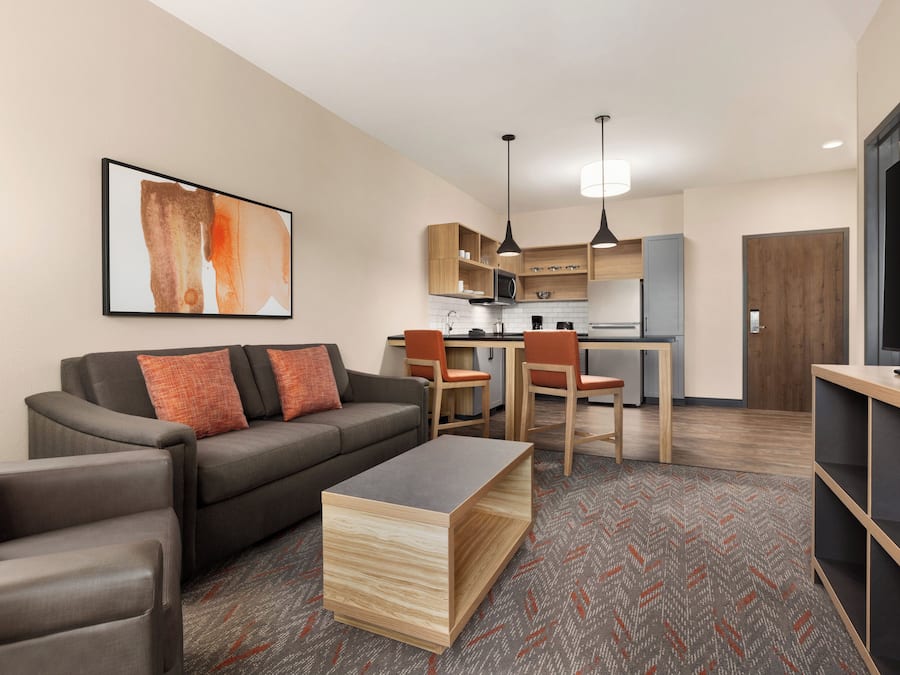 A modern living room with a sofa, coffee table, and TV stand. There is a kitchen area with cabinets and a microwave.