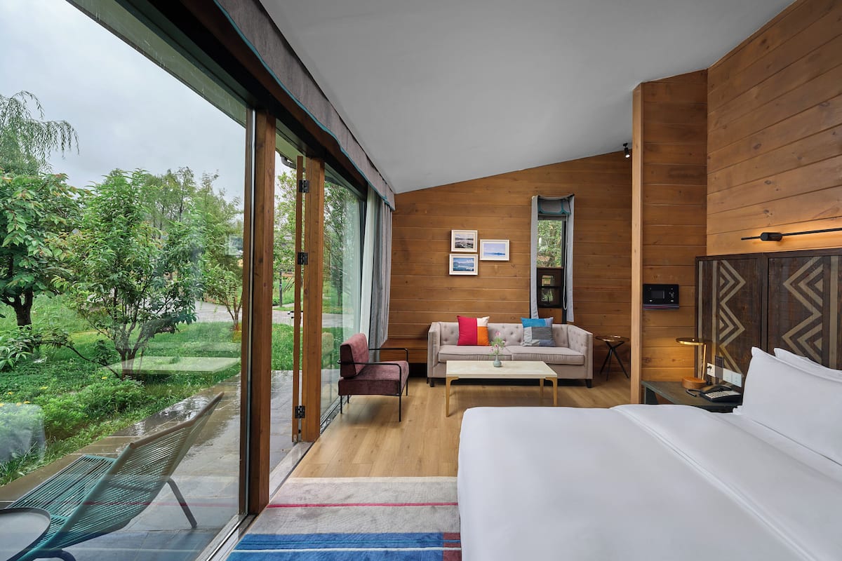 Une chambre moderne avec un grand lit, un canapé et un bureau. Une fenêtre offre une vue sur la verdure environnante.