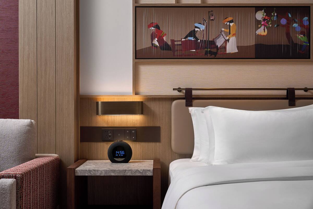Une chambre d’hôtel avec un lit, une table de chevet dotée d’une horloge numérique, un tableau au-dessus du lit et une chaise.