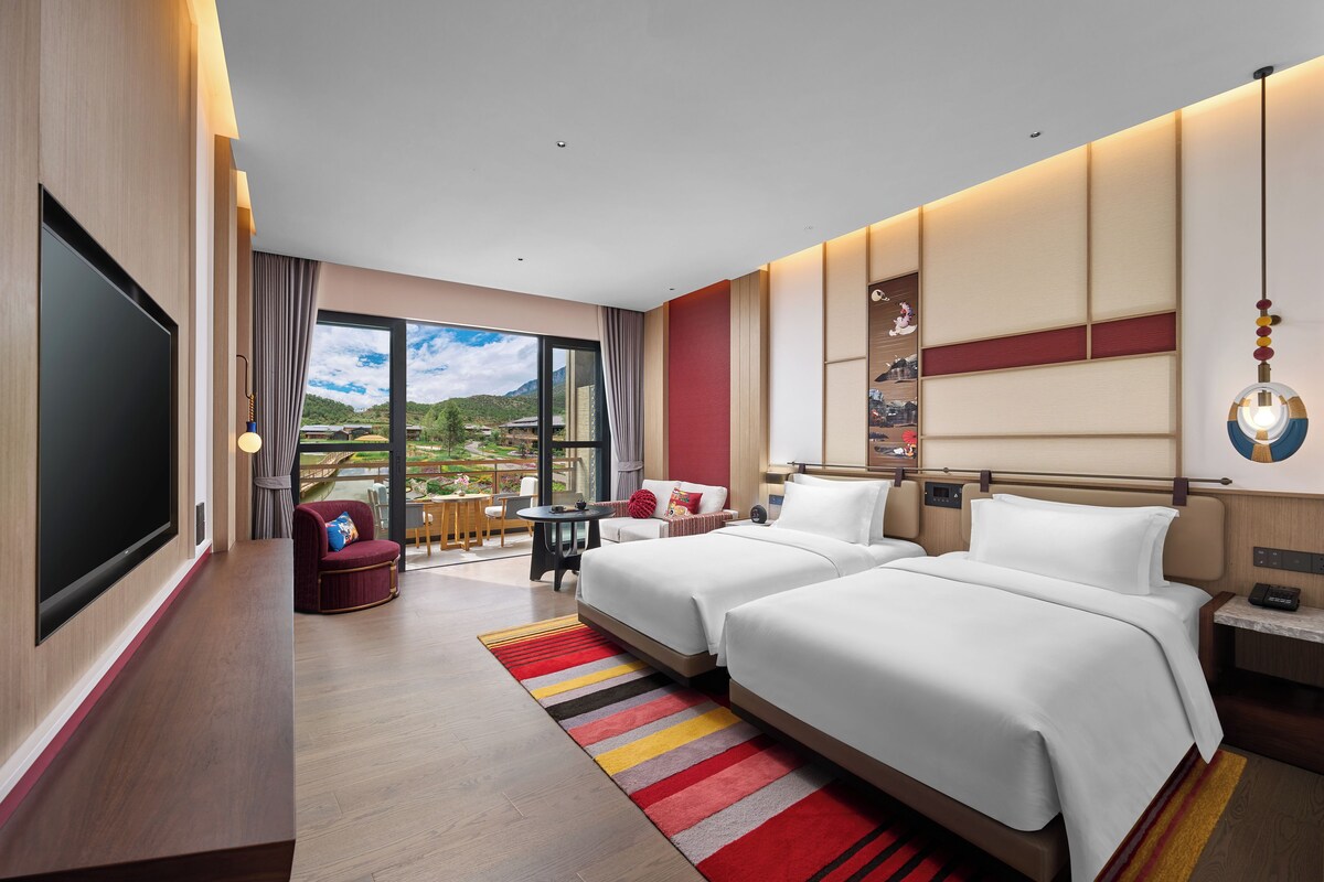 Une chambre d’hôtel avec deux lits, une télévision, un balcon donnant sur les montagnes et une petite table avec une chaise.