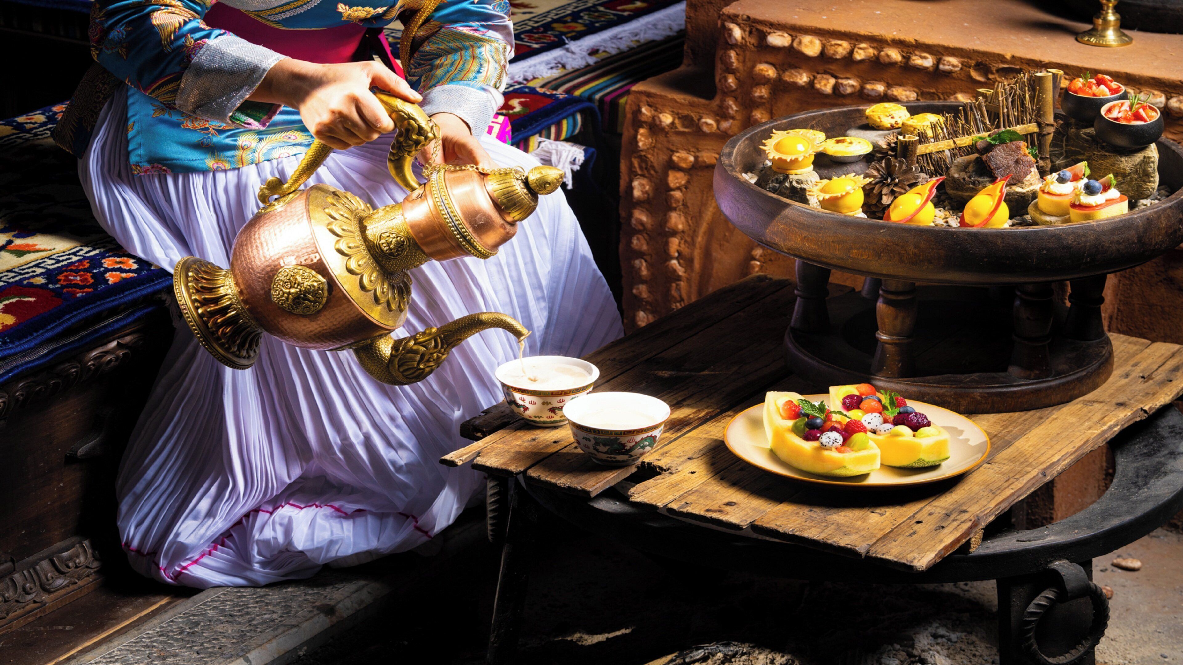 A traditional tea ceremony setup with a person pouring tea into a bowl, a fruit platter, and a decorative tea pot.