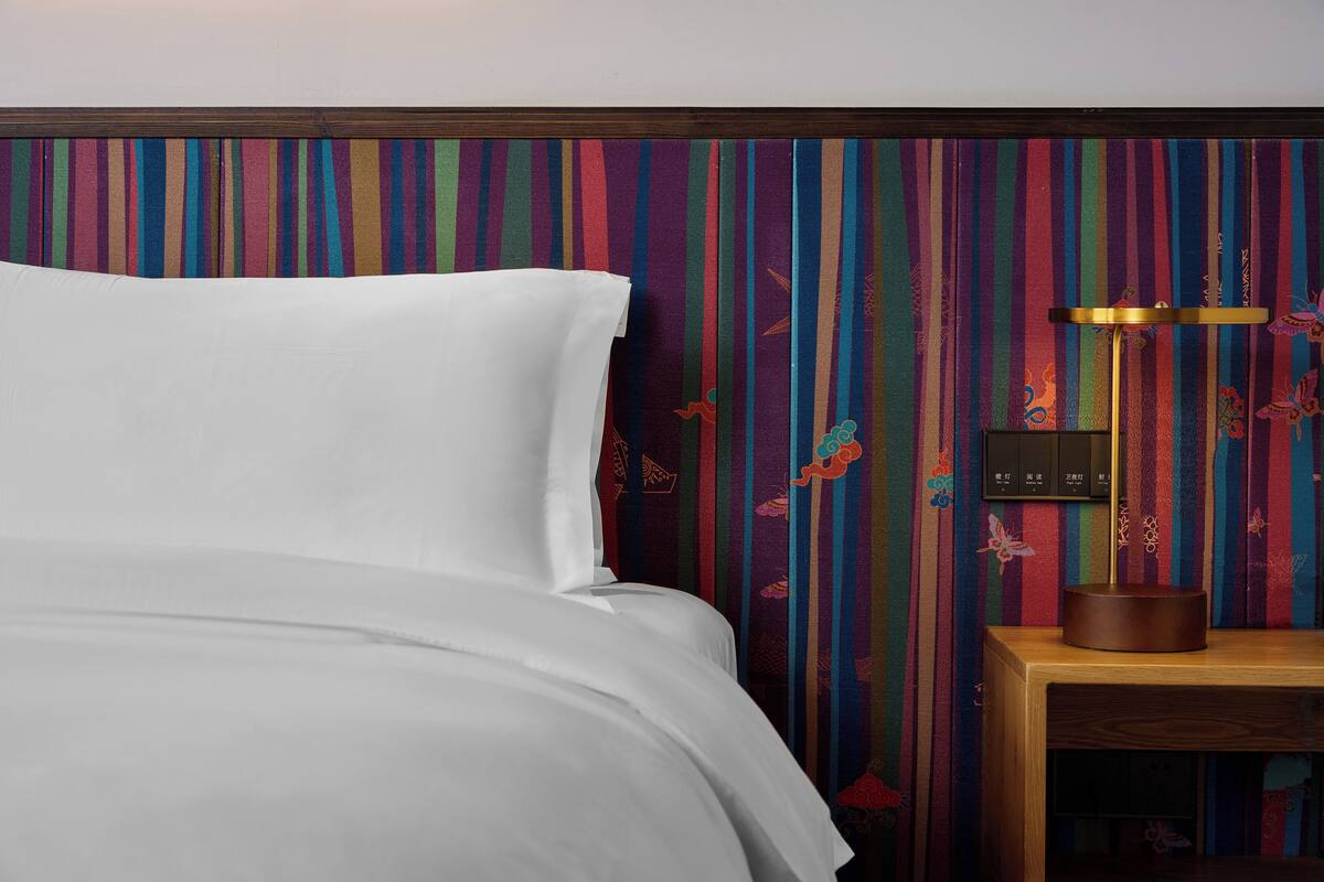 Un lit avec du linge de lit blanc, une table de chevet en bois surmontée d’une petite table ronde, et un mur à rayures colorées.