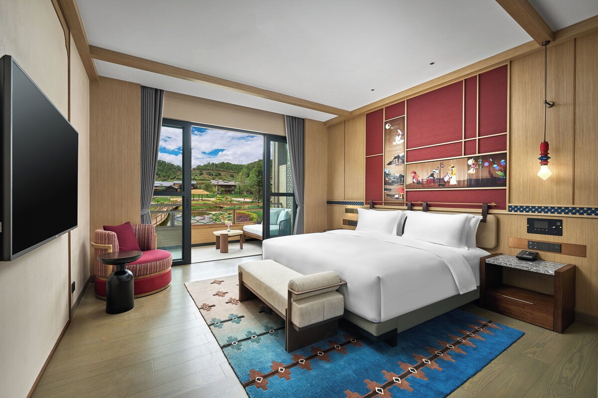 Une chambre d’hôtel moderne dotée d’un grand lit, d’une télévision et d’un coin salon avec un canapé rouge, offrant une vue sur un paysage pittoresque par la fenêtre.