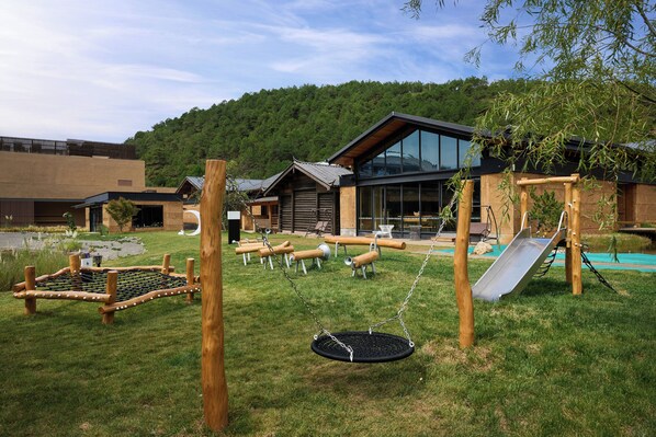 A playground with swings and a slide, surrounded by a grassy area and a building with large windows.