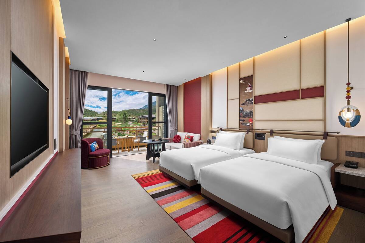 Une chambre d’hôtel avec deux lits, une télévision, un balcon donnant sur les montagnes et une petite table avec une chaise.