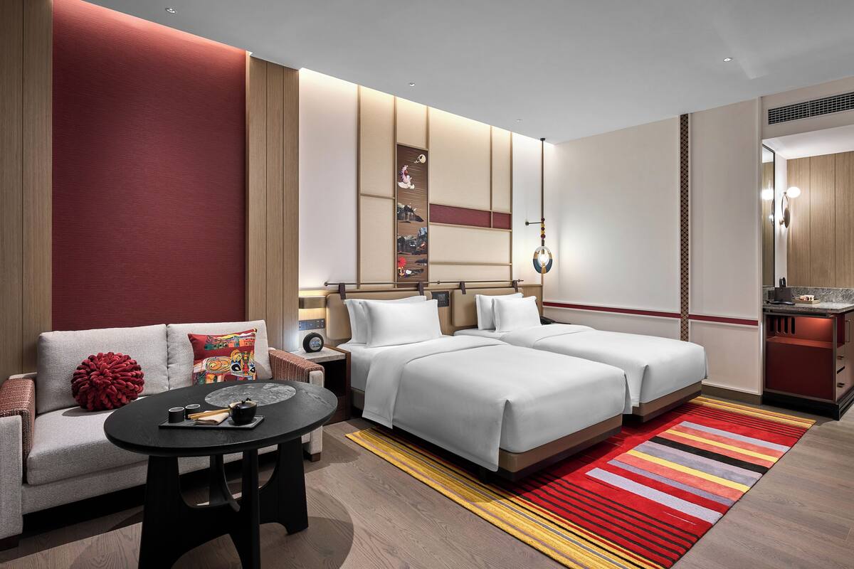 Une chambre d’hôtel avec deux lits, un canapé, une petite table avec une théière, et un tapis rayé rouge et jaune.