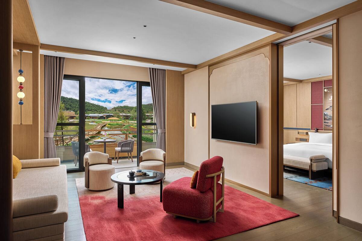 Une chambre d’hôtel avec une grande fenêtre, un téléviseur à écran plat, un canapé, un fauteuil rouge et un lit.
