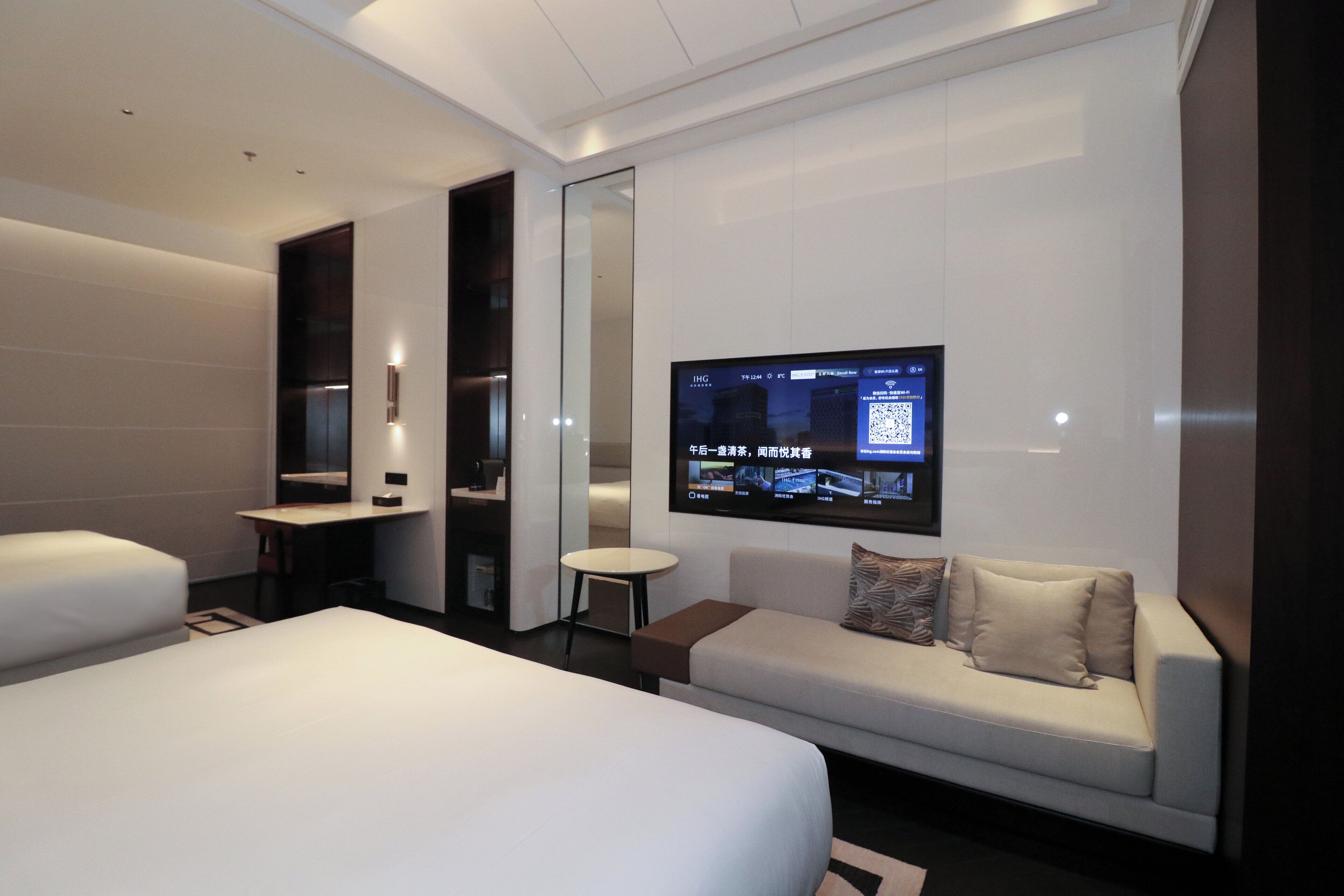 A modern hotel room with a large bed, a sofa, a small table, and a TV mounted on the wall displaying a QR code.