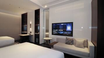 A modern hotel room with a large bed, a sofa, a small table, and a TV mounted on the wall displaying a QR code.