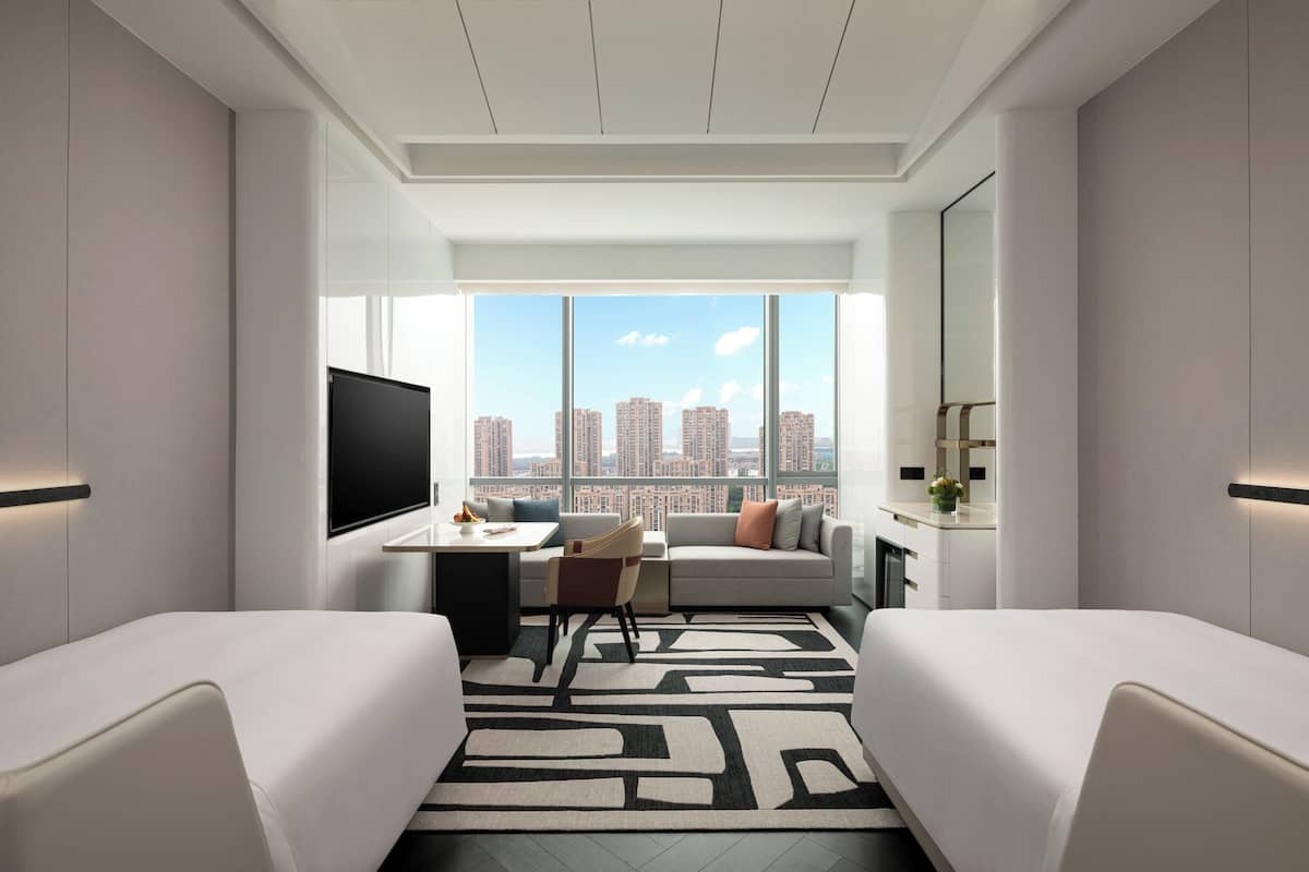 A modern hotel room with a city view, a flat-screen TV, a desk with a chair, and two beds.