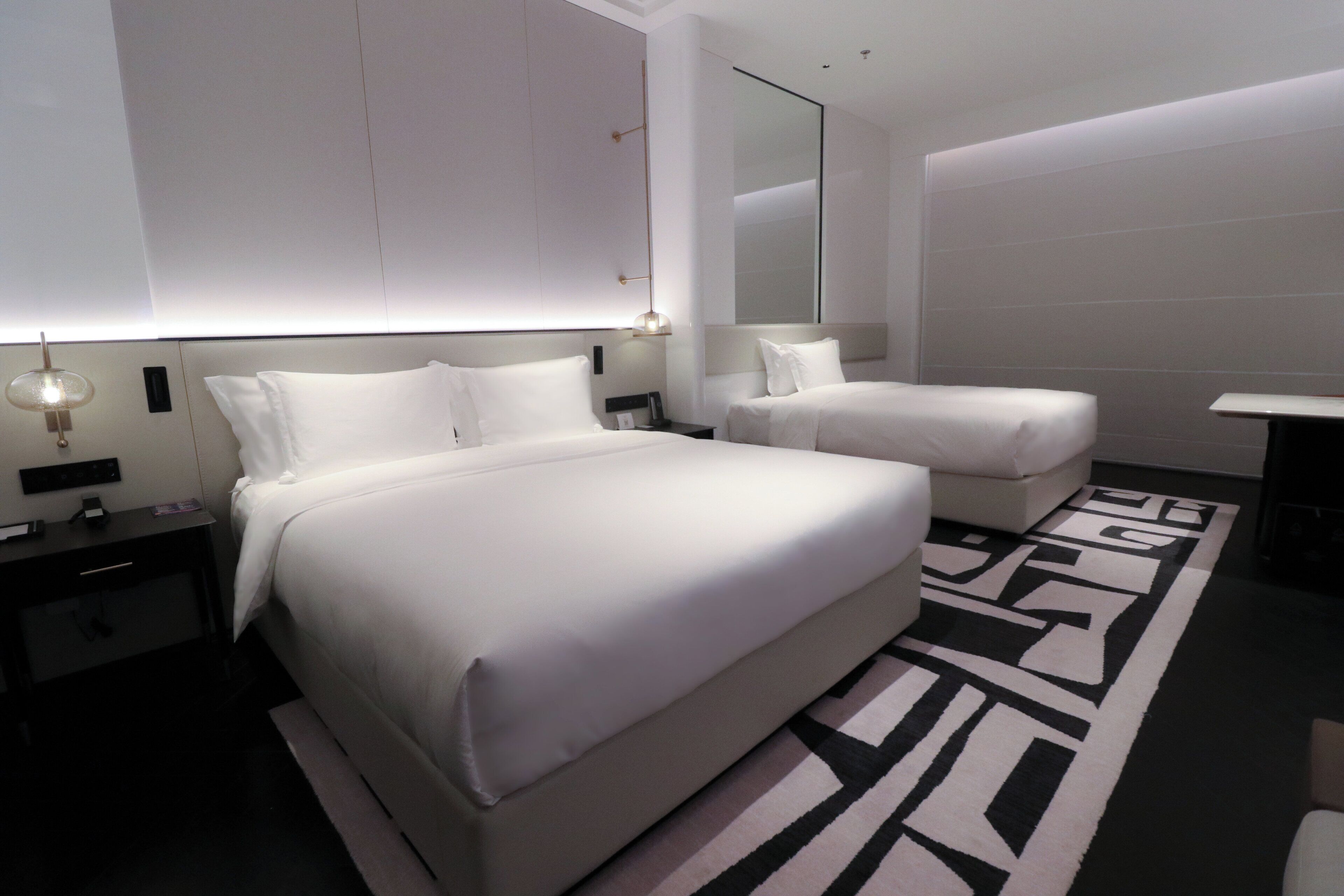 A modern hotel room with a large bed, a smaller bed, a desk, and a mirror.