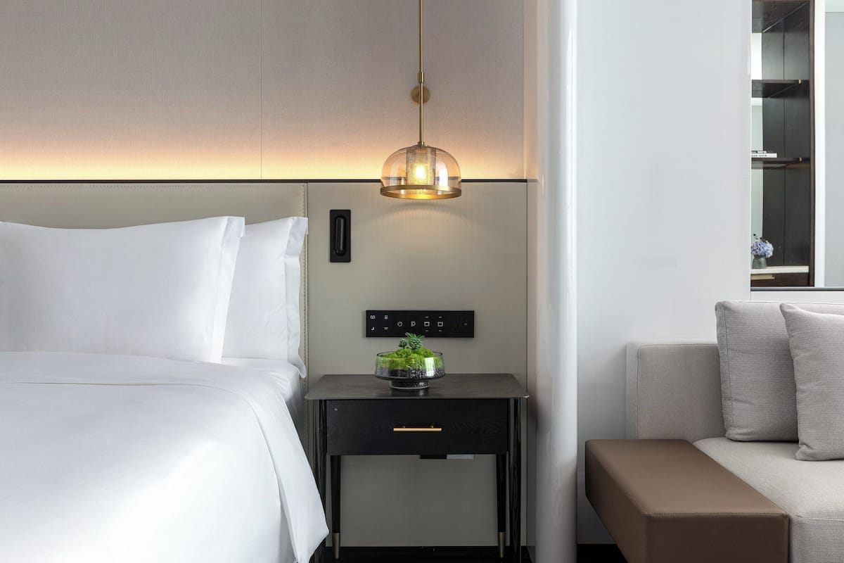 A modern hotel room with a bed, a bedside table with a lamp, a control panel, and a sofa.