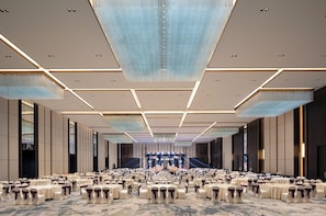 Un gran salón de banquetes con filas de mesas redondas, cada una con mantel blanco y fajas moradas. La sala tiene un techo alto con iluminación empotrada y un candelabro central.