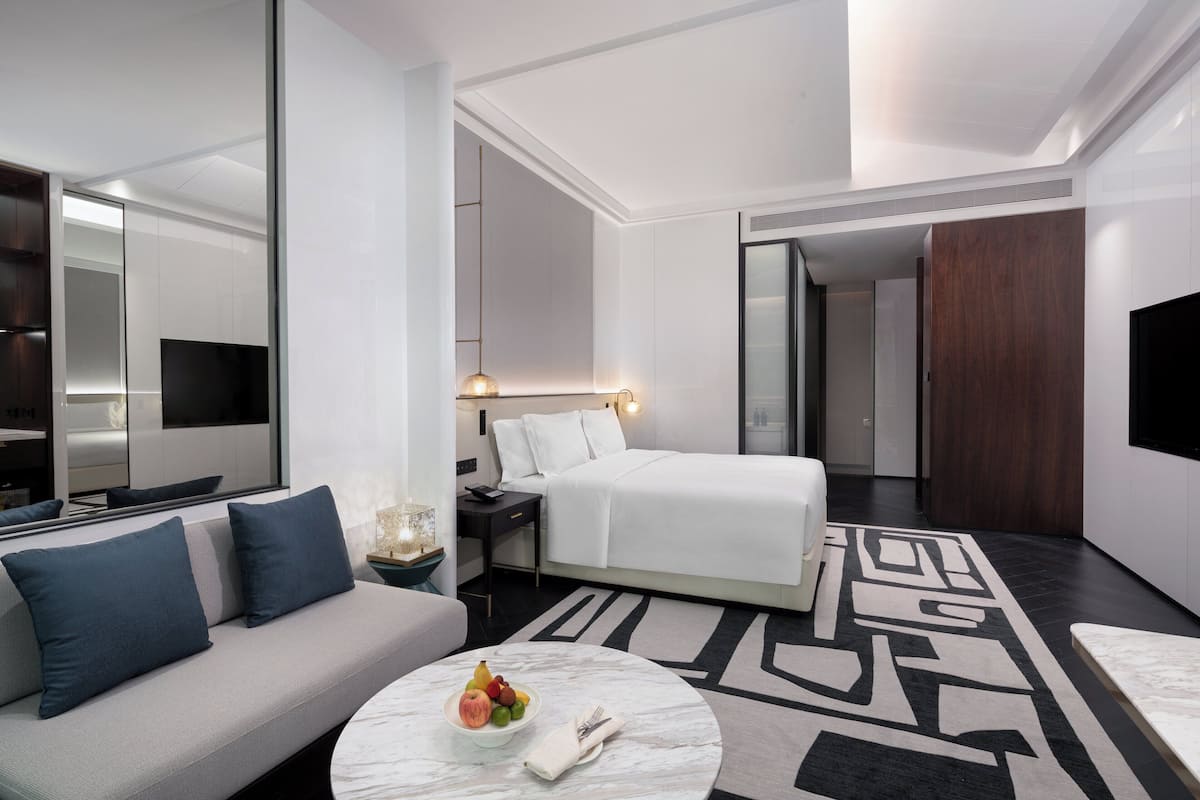 A modern hotel room with a bed, a sofa, a round coffee table with fruit, and a large mirror.