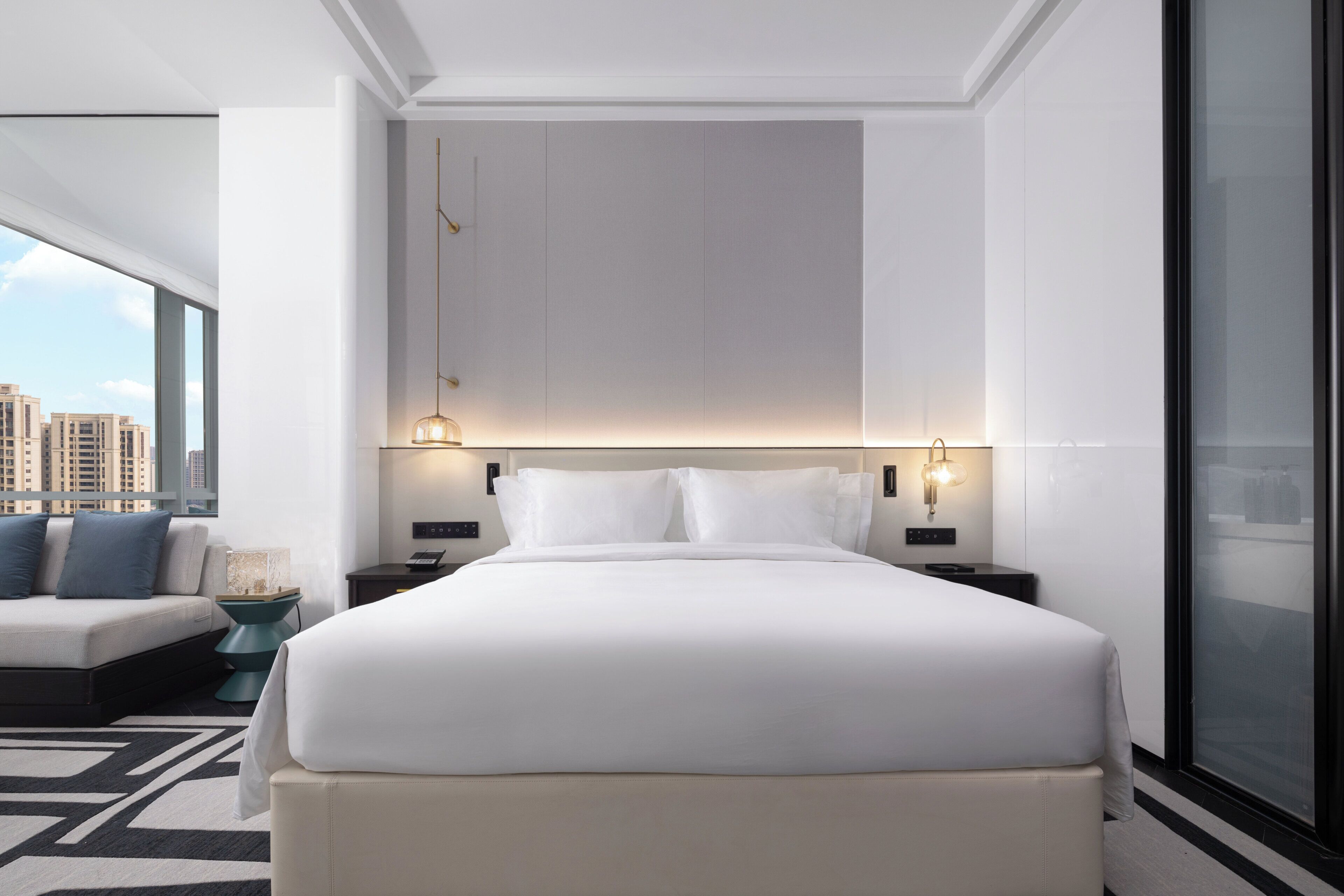 A modern hotel room with a large bed, bedside tables, a nightstand, a lamp, and a view of the city through the window.