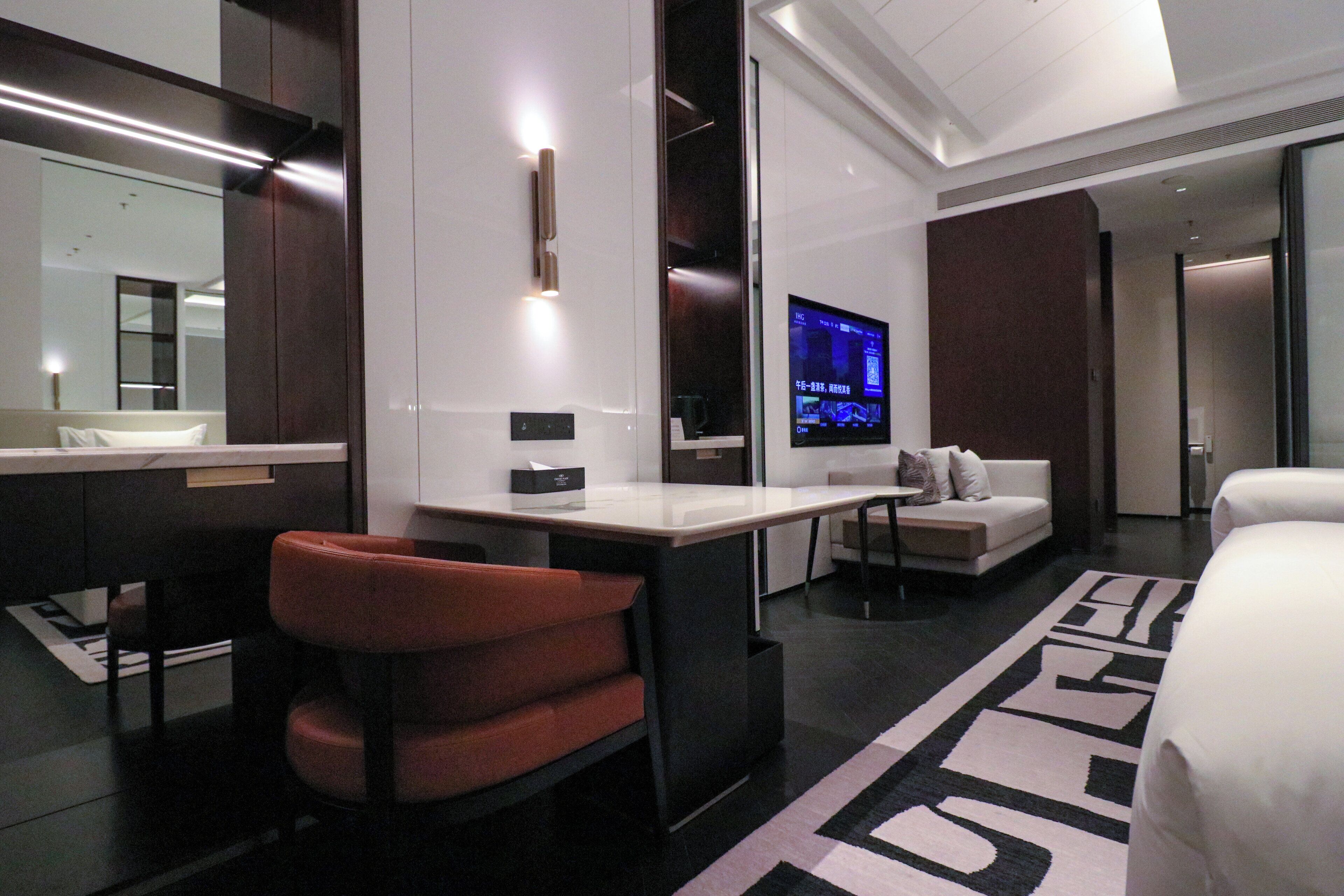 A modern hotel room with a large bed, a desk, a chair, a TV, and a bathroom with a mirror and sink.