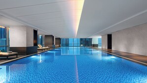 Una piscina interior moderna con suelo de baldosas azules, sillas de descanso y un muro de cristal en un lateral.